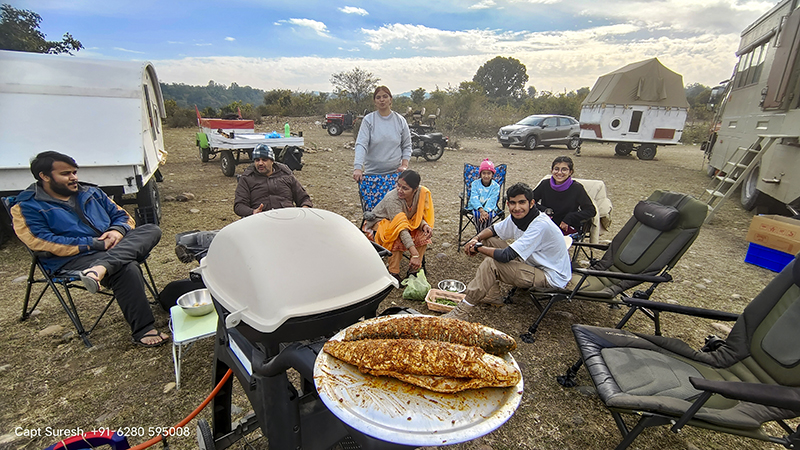 family of guests enjoying freshly cooked fish in a weber barbecue grill q3200 at offbeat location around pong dam in himachal pradesh with caravan in wilderness. 